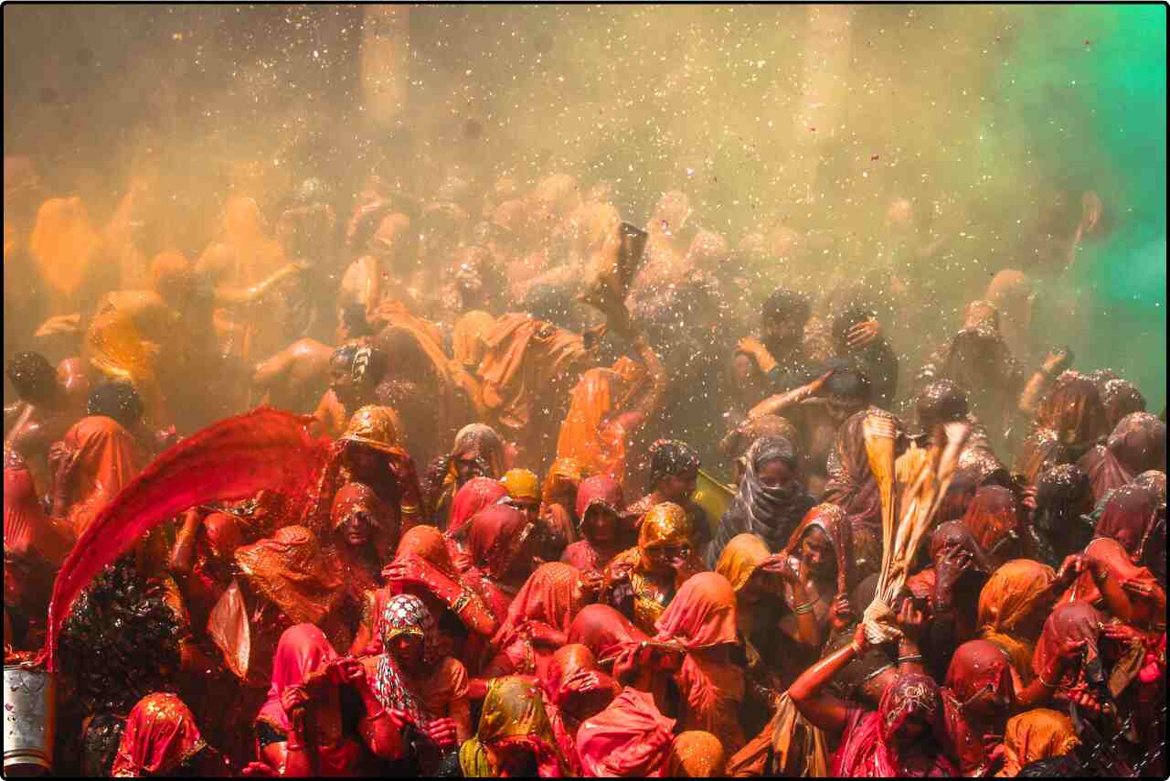 Joyful crowd in red shirts celebrating Holi, with hands raised and covered in colorful powder.